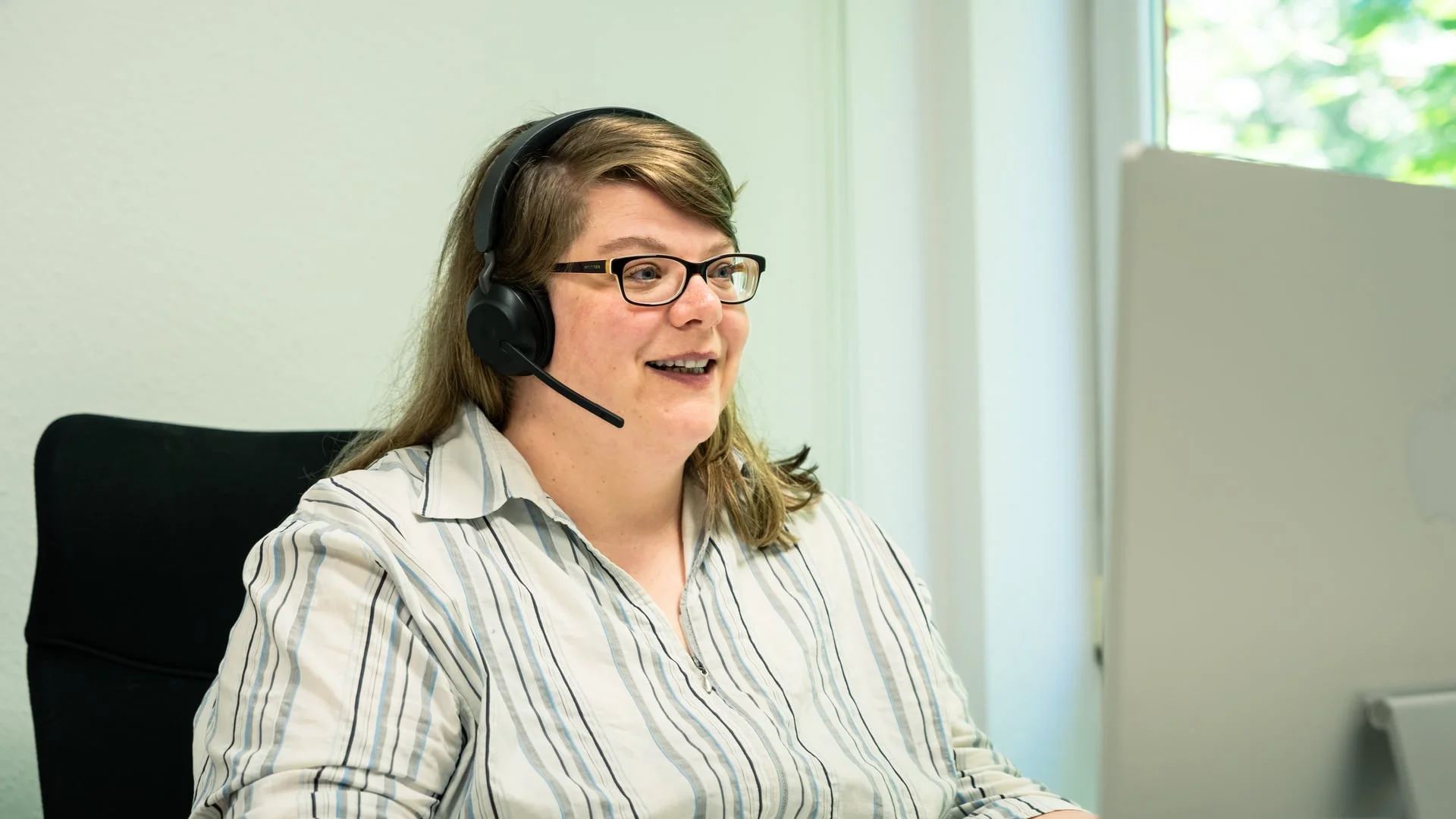 Frau mit Brille und Headset lächelt bei der Arbeit am Bildschirm im hellen Büro