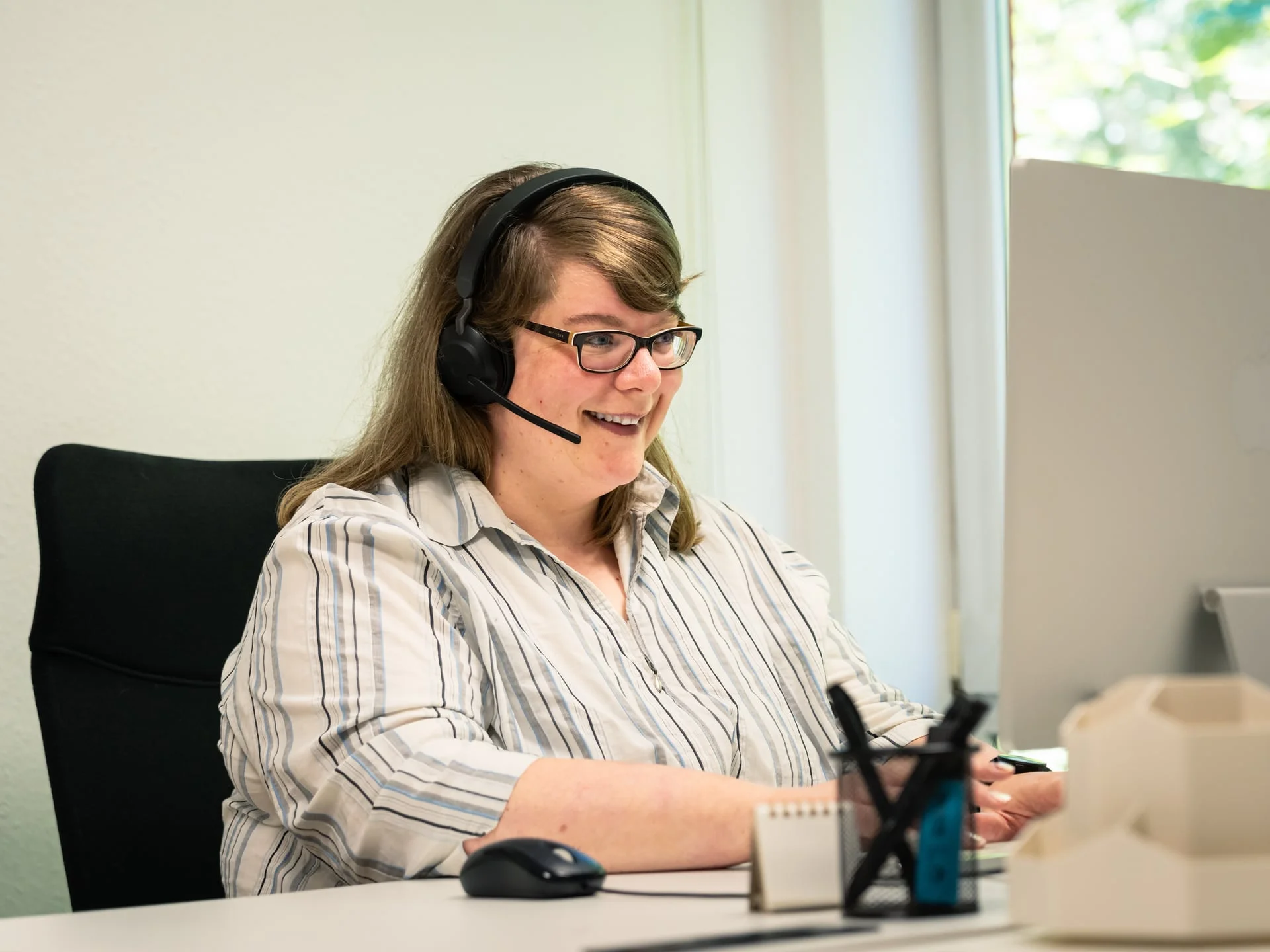 Frau mit Headset und Brille lächelt am Schreibtisch im hellen Büro