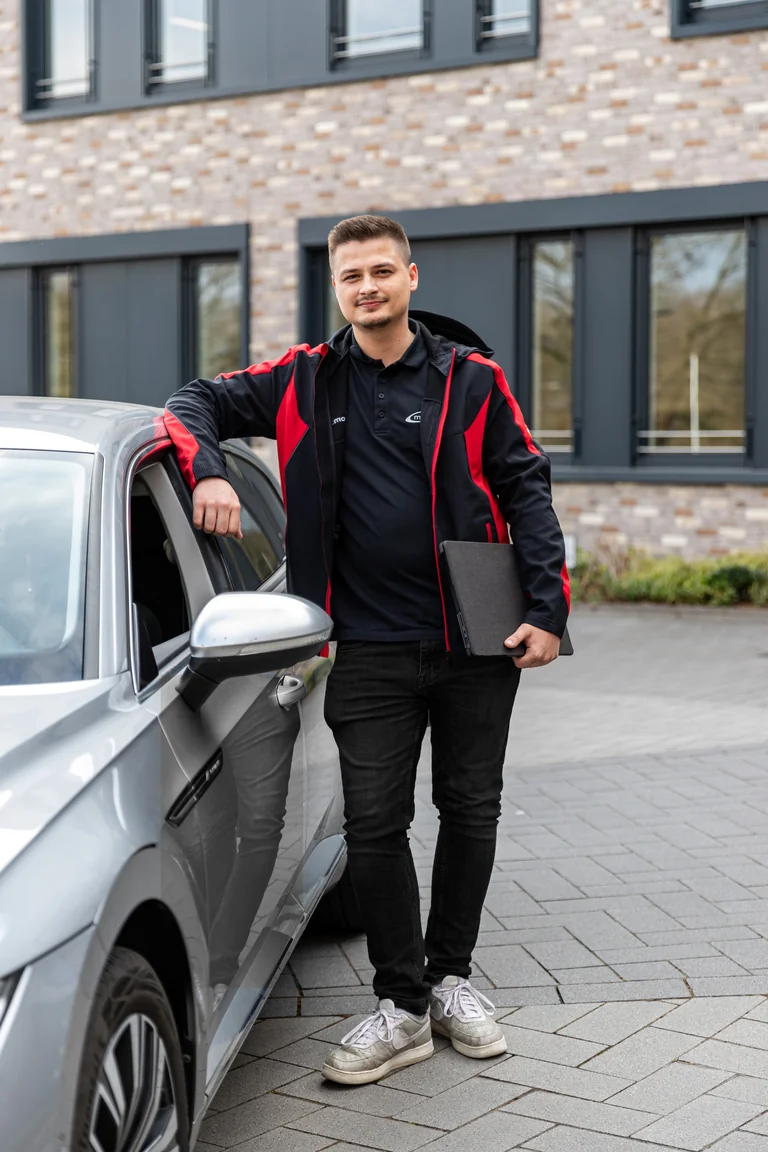 Manuel Neitzel, Support & Technik bei medacom, in roter Jacke mit Notebook am Auto vor dem Firmengebäude