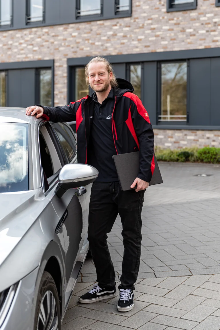 Patrick Oelrich, Leitung Support & Technik bei medacom, in rot-schwarzer Jacke mit Notebook am silbernen Auto